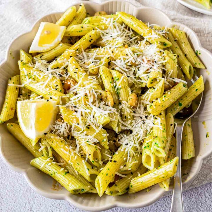 A bowl of penne pasta coated in a light green herb sauce, topped with shredded cheese, small shrimp, and black sesame seeds, garnished with lemon wedges.
