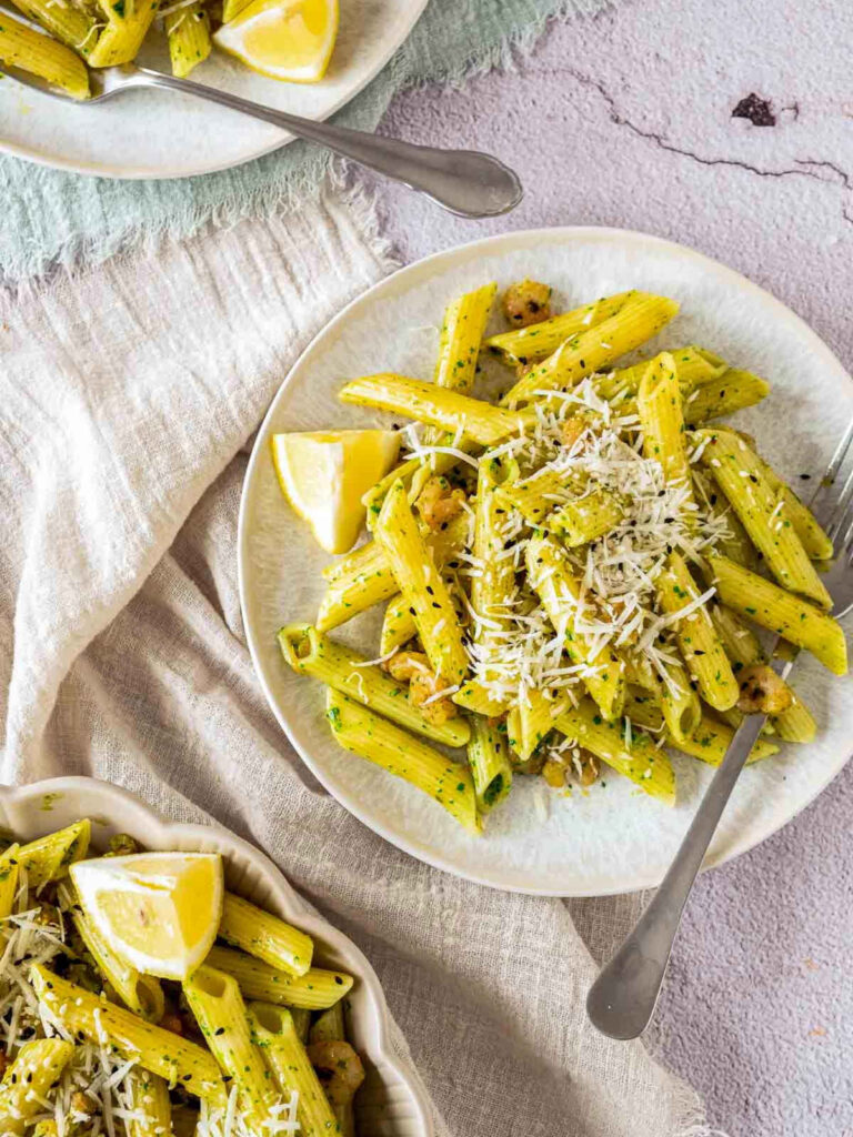 A top-down view of three plates of penne pasta tossed in a vibrant green herb or pesto sauce. Each portion is garnished with a generous mound of shredded parmesan cheese and small cooked shrimp. A fresh lemon wedge sits on the side of each plate, and the dishes are set on a rustic, light-colored linen cloth over a textured grey stone surface. Stainless steel forks are placed beside the plates.