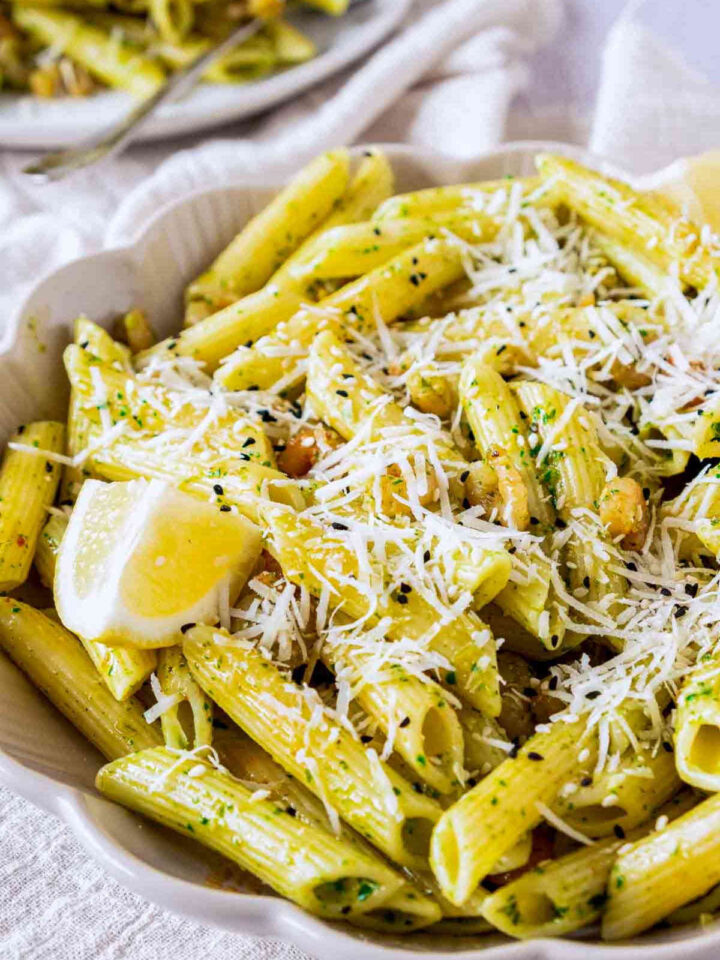 A close-up of penne pasta tossed in a vibrant green pesto sauce, topped with shredded parmesan cheese, small shrimp, and black sesame seeds, served with a fresh lemon wedge in a white scalloped bowl.