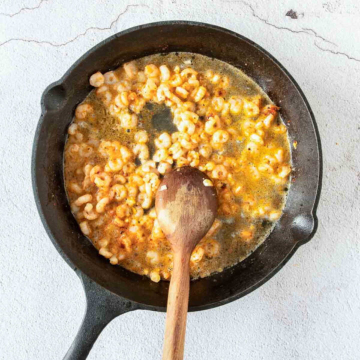 A wooden spoon stirs small, pink shrimp cooking in a bubbling garlic butter sauce within a black cast-iron skillet.