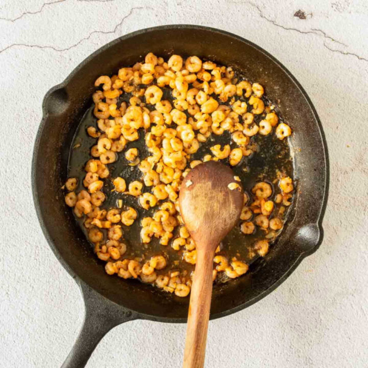 Cooked, golden-brown shrimp being stirred with a wooden spoon in a cast-iron skillet after the sauce has reduced.