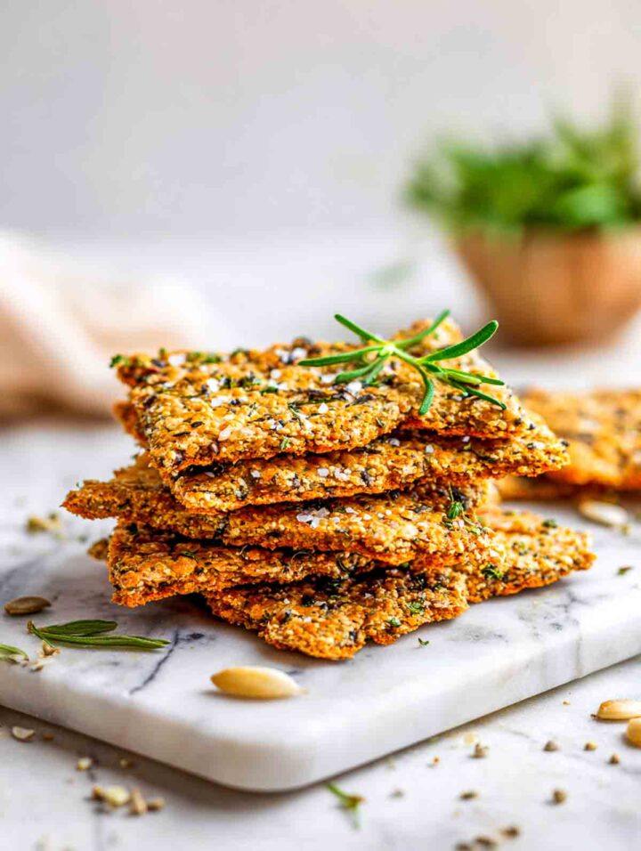 Crunchy green lentil flatbread crackers stacked on marble surface with herbs in the background.