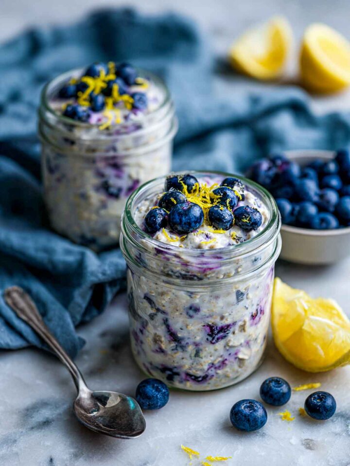 Two glass jars of lemon blueberry overnight oats filled close to the rim, with blueberries mixed throughout and on top, finished with fresh lemon zest, styled on a white marble surface with lemon wedges, a blue-gray linen napkin, and a spoon.