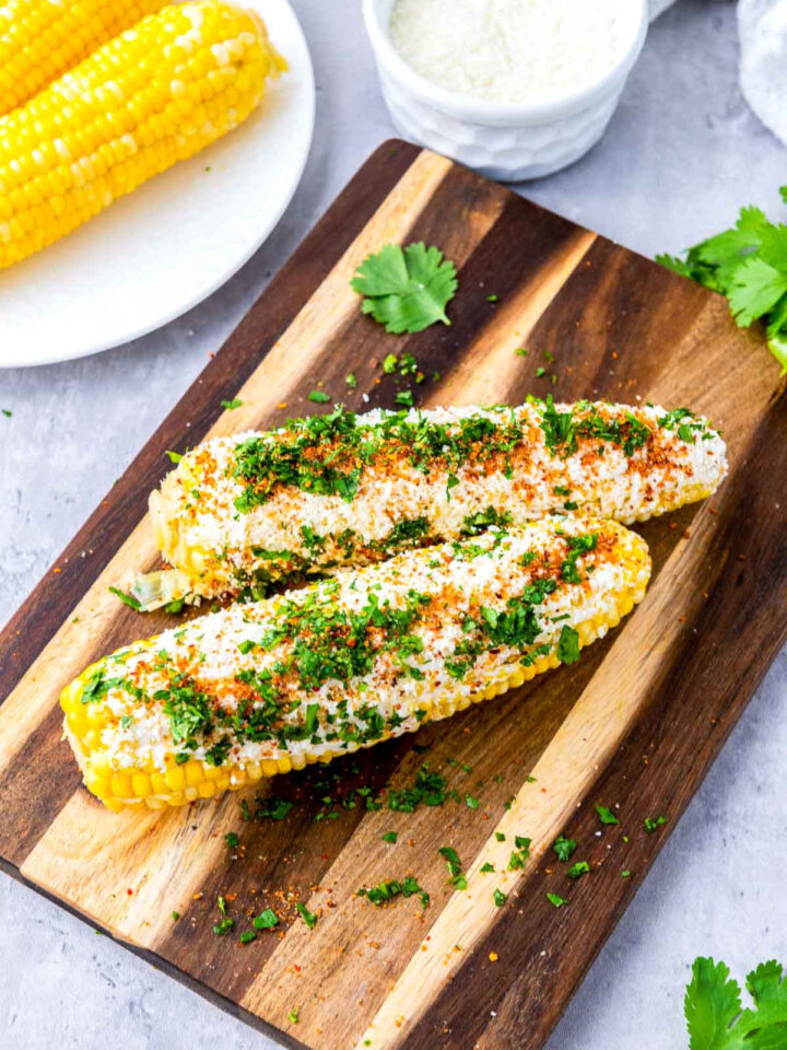Mexican-style elotes on a wooden cutting board, coated with creamy cheese, chopped cilantro, and chili seasoning, styled on a light kitchen surface with extra corn and grated cheese in the background.
