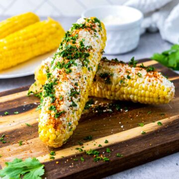 Mexican street corn (elotes) resting on a wooden board, generously coated with creamy cheese, chili powder, and fresh cilantro, with extra corn cobs and a small bowl of sauce softly blurred in the background.