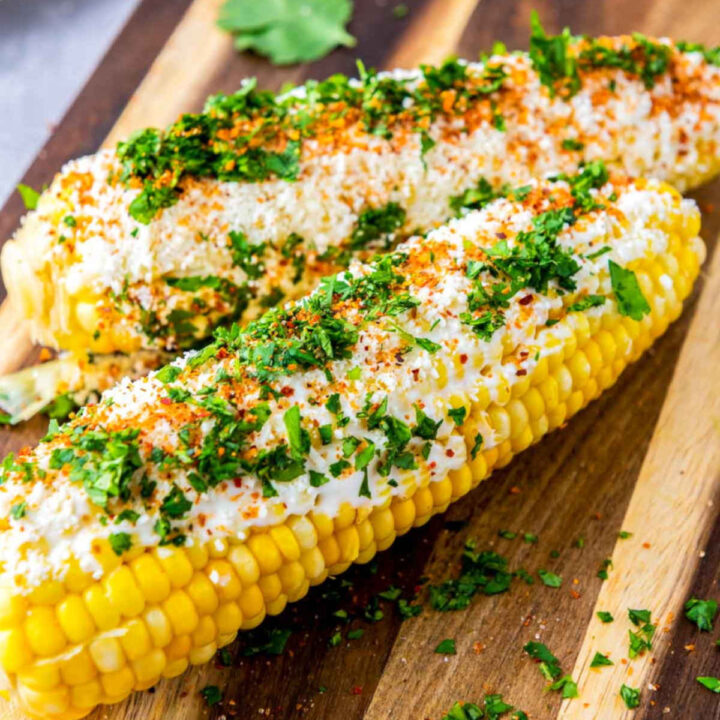 Close-up of Mexican street corn (elotes) coated in creamy sauce, crumbled cheese, chili seasoning, and fresh cilantro, served on a wooden cutting board with scattered herbs.