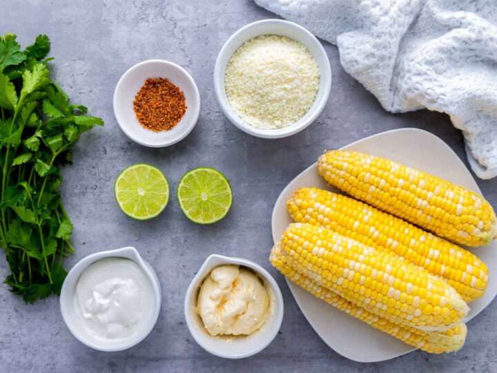 Ingredients for Mexican street corn (elotes) arranged on a gray surface: fresh corn on the cob, crumbled cheese, mayonnaise, sour cream, chili seasoning, halved limes, and fresh cilantro, with a textured white cloth nearby for a casual kitchen feel.