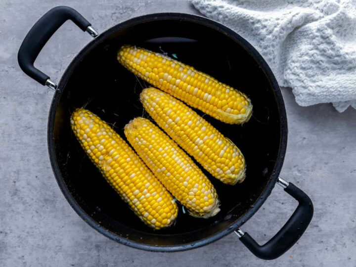 Four ears of yellow corn boiling in a large black pot filled with water, viewed from above on a gray countertop, with a white kitchen towel nearby for a simple, everyday cooking scene.