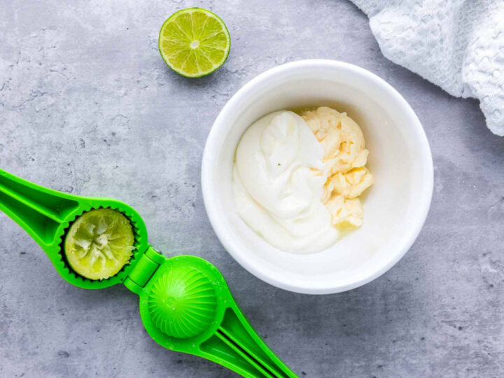 A white bowl with mayonnaise and softened butter beside a bright green lime squeezer and a halved lime on a gray countertop, showing the creamy base being prepared for elotes sauce.