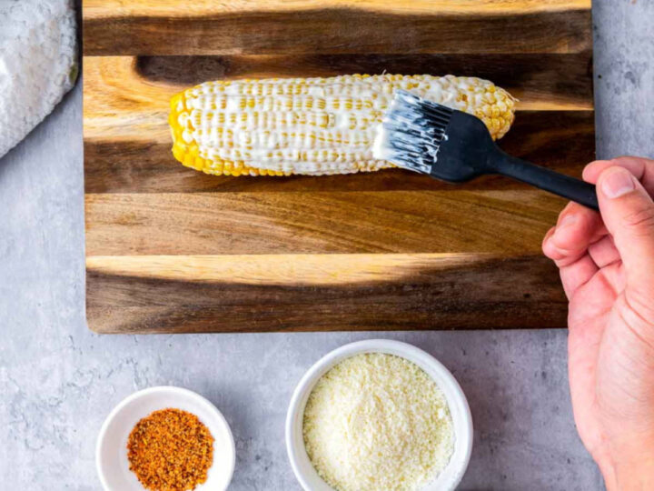 A hand uses a silicone brush to coat a cooked ear of corn with a creamy sauce on a wooden cutting board, with bowls of cheese and seasoning nearby, showing the elotes being assembled.