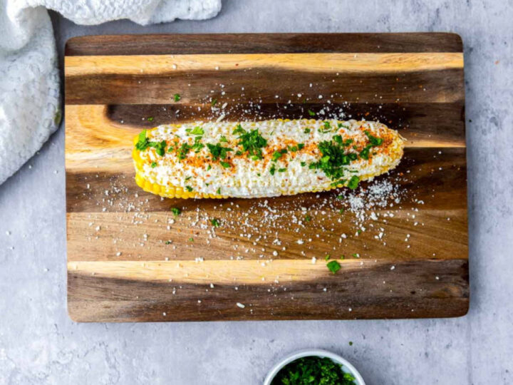 Finished Mexican street corn (elotes) topped with creamy sauce, crumbled cheese, chili seasoning, and chopped cilantro, resting on a wooden cutting board with scattered cheese crumbs.