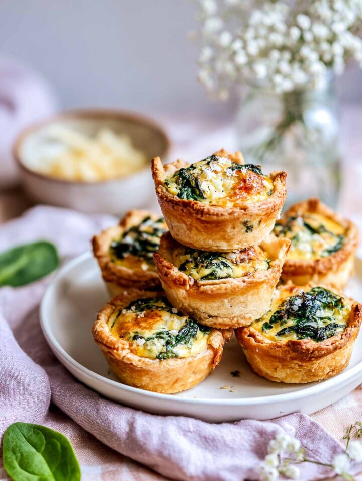 Mini spinach and cheese quiche bites stacked on a white plate with flowers in the background.