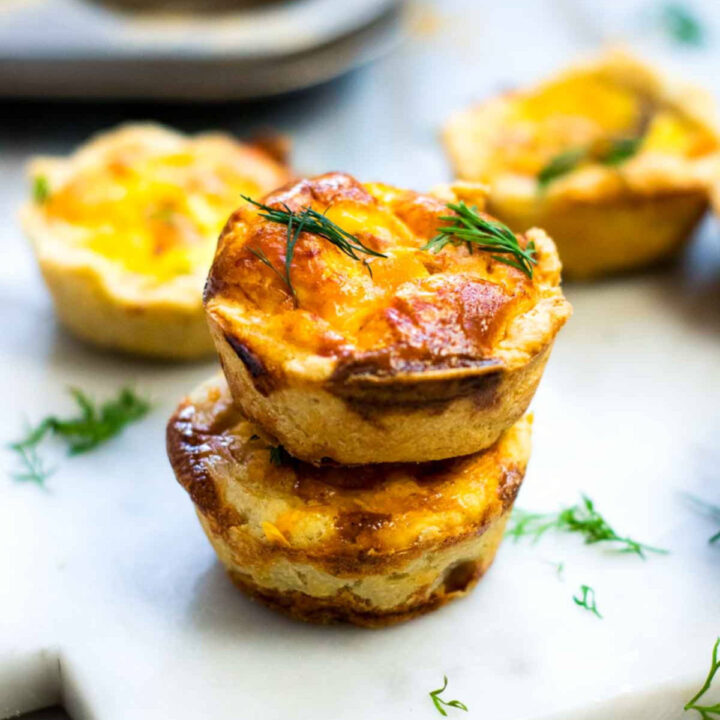 Stacked mini salmon quiches with golden, flaky crusts and lightly browned tops, garnished with fresh dill on a white serving surface.