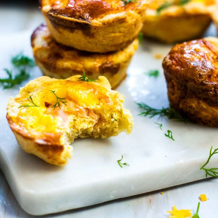 Close-up of mini salmon quiches on a white board, one cut open to show a creamy egg and salmon filling, with a golden-brown crust and fresh dill scattered around.