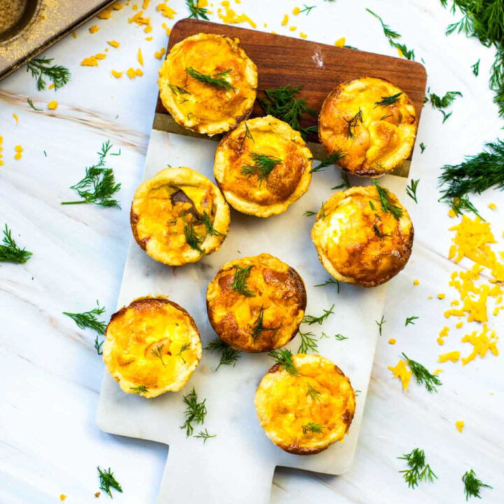 Overhead view of mini salmon quiches with golden, lightly browned tops arranged on a marble board, garnished with fresh dill and scattered shredded cheese.