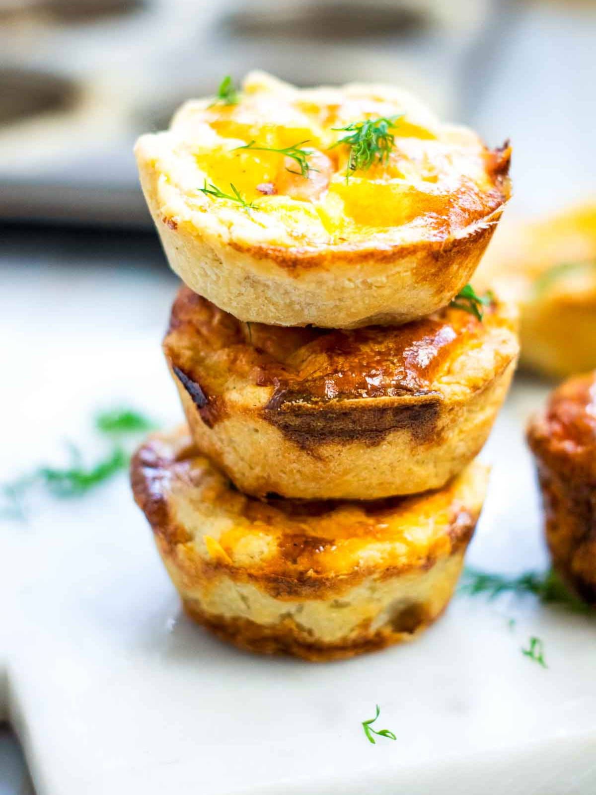 Three stacked mini salmon quiches with golden, flaky crusts and lightly browned tops, garnished with fresh dill on a white serving surface.