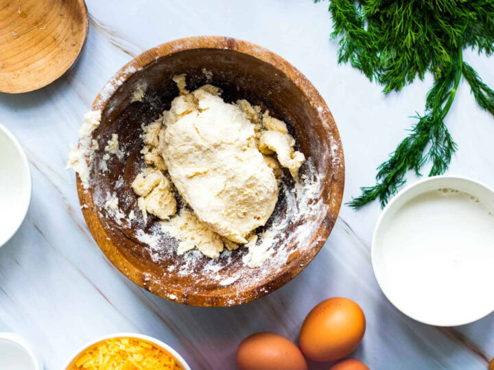 Top-down view of quiche dough partially mixed in a wooden bowl, with flour-dusted sides, surrounded by eggs, milk, shredded cheese, and fresh dill on a light marble surface.