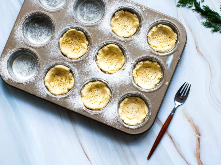 Top-down view of a floured muffin tin with several cups lined with pressed quiche pastry shells, docked with fork holes, resting on a marble surface with a fork beside it.