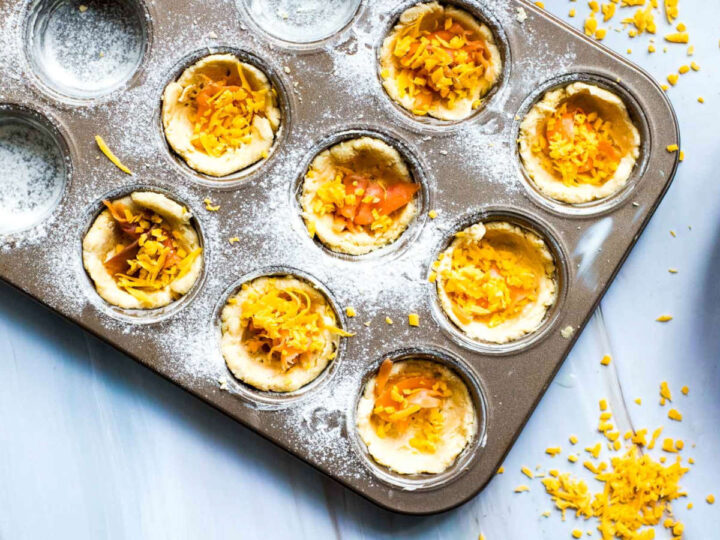 Overhead view of a muffin tin with mini quiche pastry shells filled with shredded cheese and small pieces of salmon, lightly dusted with flour, with extra shredded cheese scattered on a light marble surface beside the pan.