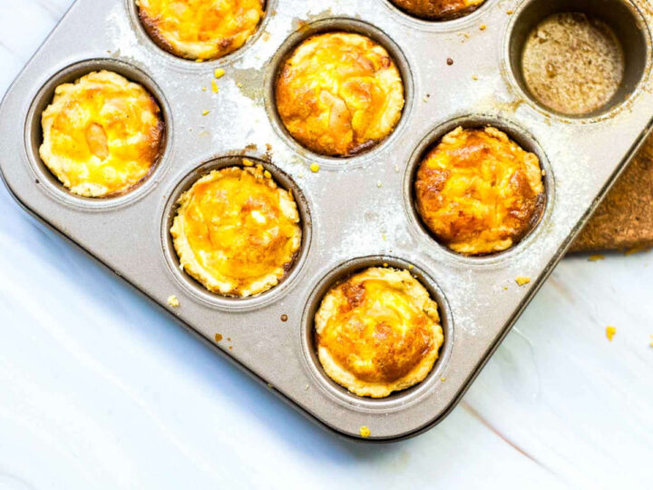 Overhead view of a muffin tin filled with freshly baked mini salmon quiches, golden and slightly puffed with melted cheese on top, set in crisp pastry shells with a few crumbs scattered around the pan.
