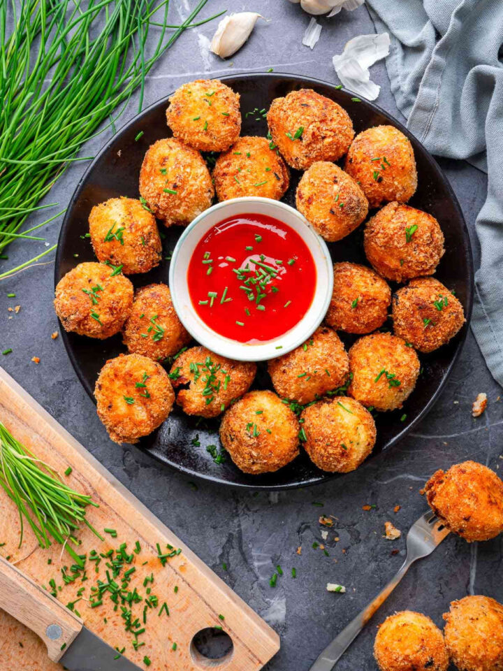 Crispy parmesan herb potato bites arranged on a black plate around a bowl of red dipping sauce, garnished with fresh chives on a rustic surface.