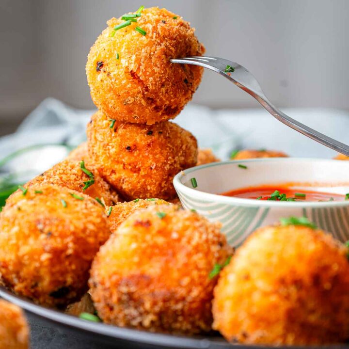 Golden, crispy potato bites stacked on a plate, topped with chopped chives, with one bite lifted on a fork beside a bowl of red dipping sauce.