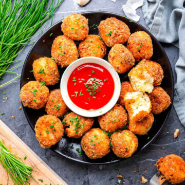 Crispy parmesan herb potato bites arranged in a circle on a black plate with a bowl of red dipping sauce in the center, garnished with chopped chives; one bite broken open to show the creamy potato interior.