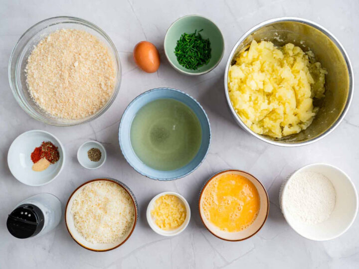 Overhead view of ingredients for parmesan herb potato bites arranged on a countertop, including mashed potatoes, breadcrumbs, flour, grated cheese, eggs, chopped herbs, seasonings, and oil in small bowls.