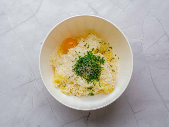 Mashed potatoes in a mixing bowl topped with a cracked egg, grated parmesan cheese, chopped fresh herbs, and seasoning before mixing.