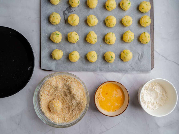 Prepared potato balls arranged on a parchment-lined baking tray, with bowls of flour, beaten egg, and breadcrumbs set up below for breading before cooking.