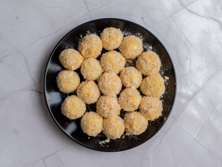 Breaded potato bites coated in breadcrumbs and arranged in a single layer on a black plate, ready to be baked or fried.