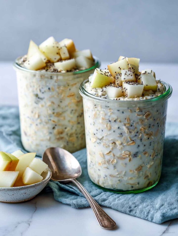 Two glass jars of vanilla pear overnight oats with chia, filled nearly to the rim and topped with diced pear and chia seeds on a white marble surface with a blue-green linen napkin and spoon.