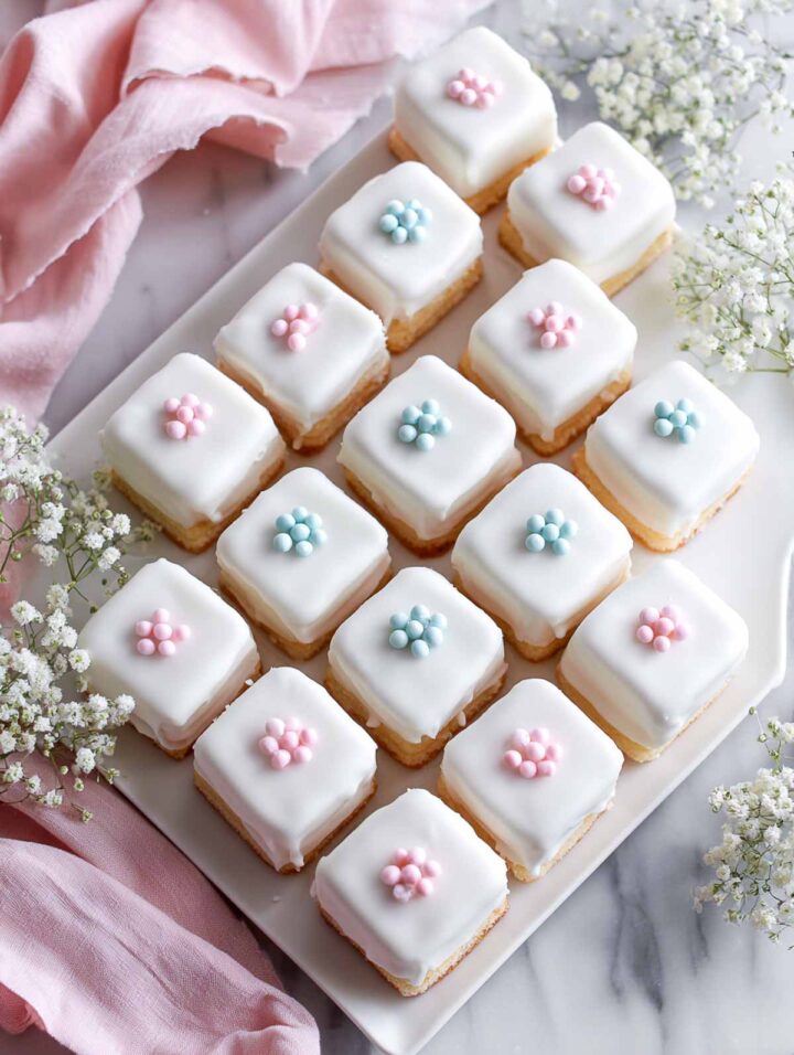 Fully glazed petit four cake squares topped with pastel sugar flowers on a white platter.