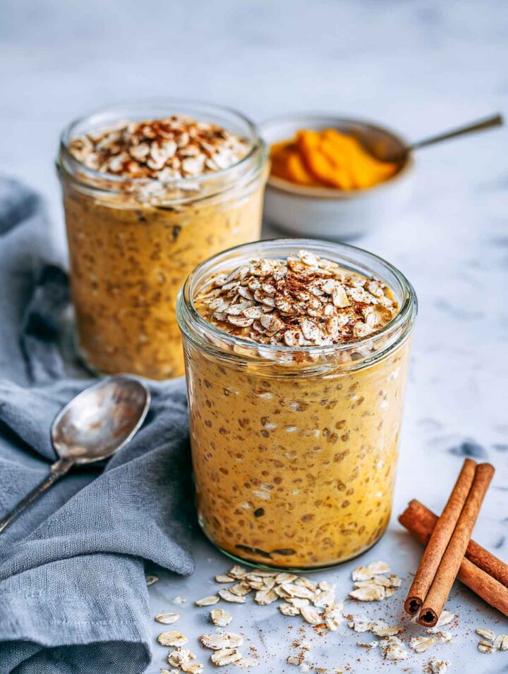Two glass jars of pumpkin spice overnight oats topped with rolled oats and cinnamon on a white marble surface with cinnamon sticks and a small bowl of pumpkin purée nearby.