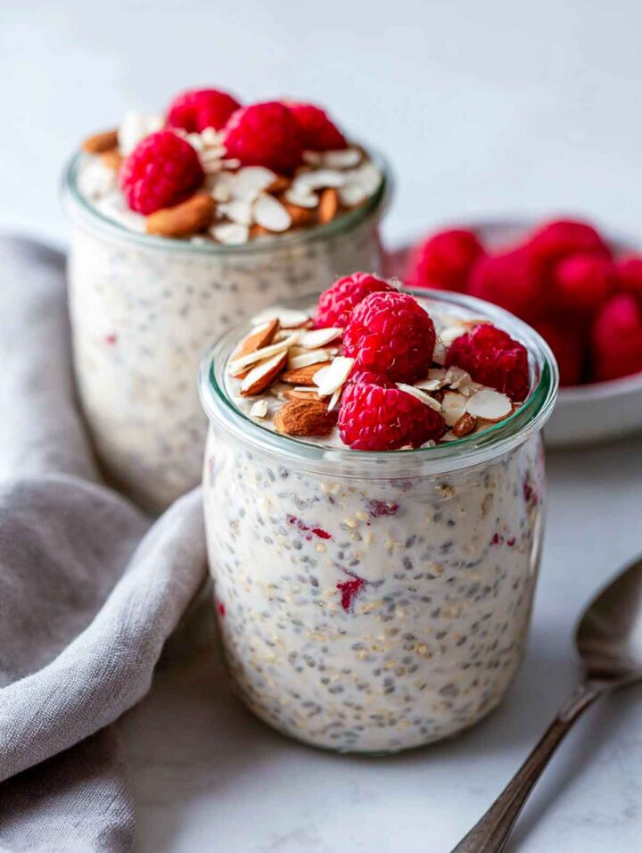 Two glass jars of raspberry almond overnight oats with chia topped with fresh raspberries and sliced almonds on a white marble surface with a neutral linen napkin.