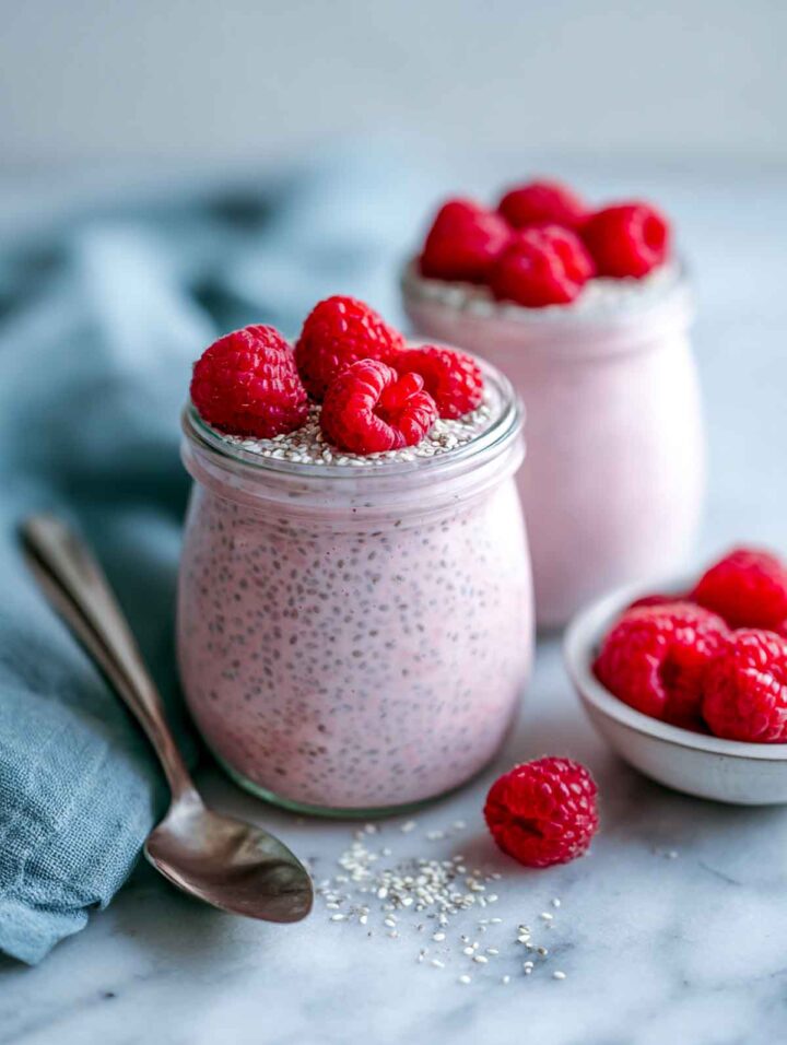 Two glass jars of raspberry kefir chia pudding filled nearly to the rim and topped with fresh raspberries on a white marble surface with a cool-toned napkin and small bowl of raspberries nearby.