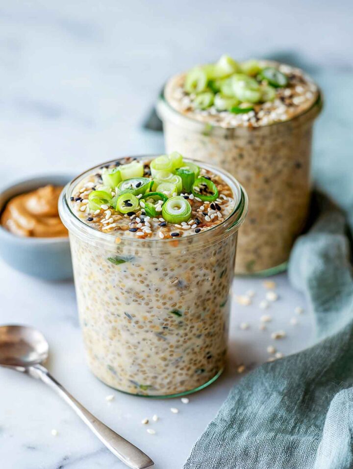 Two small glass jars filled with savory miso chia pudding mixed with oats, topped with sliced green scallions and sesame seeds, set on a white marble surface with a small bowl of miso paste and a linen napkin in the background.