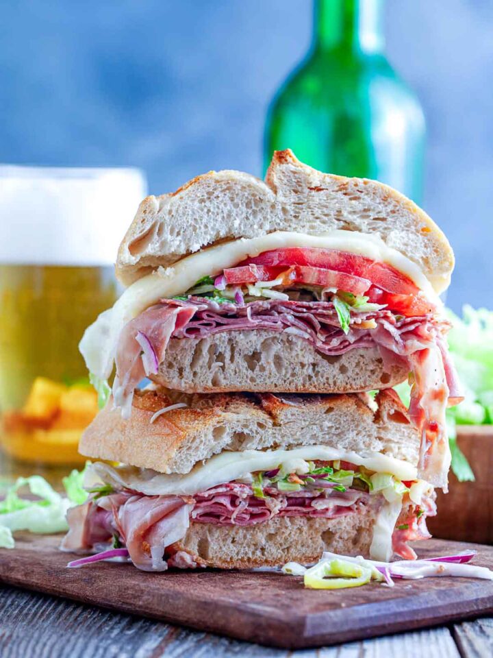 Stacked toasted Italian grinder sandwich with deli meats, melted cheese, lettuce, tomato, and onions on a cutting board.