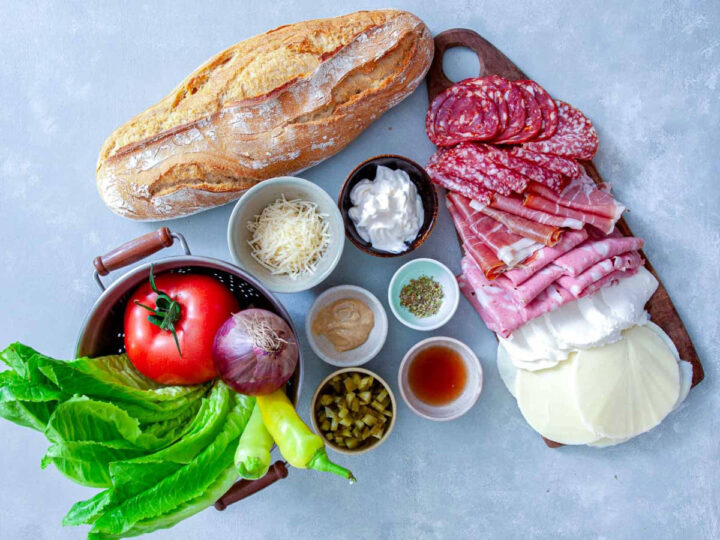 Ingredients for toasted Italian grinders including bread, deli meats, cheese, lettuce, tomato, onion, peppers, and condiments.