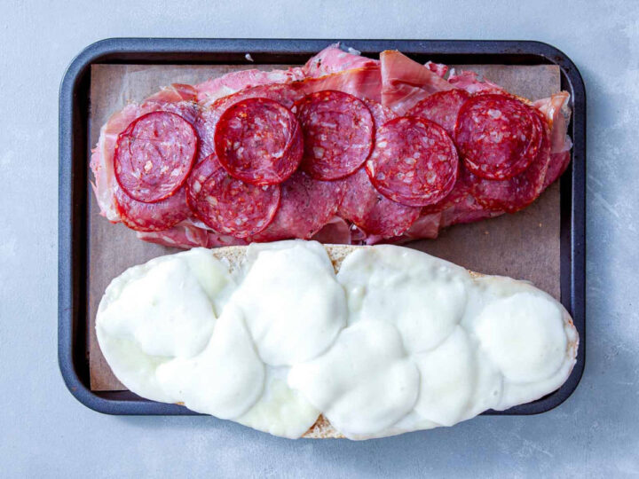 Italian grinder meats and melted cheese layered on sliced bread on a baking tray.
