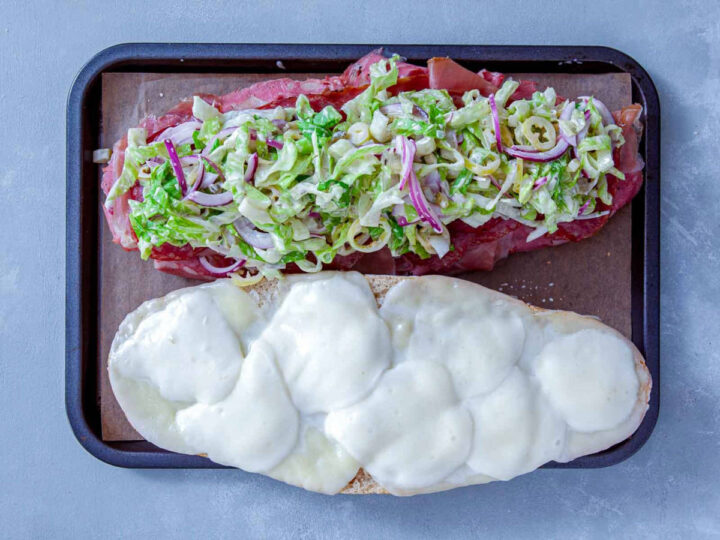 Italian grinder topped with dressed lettuce and onions over layered deli meats on a baking tray.