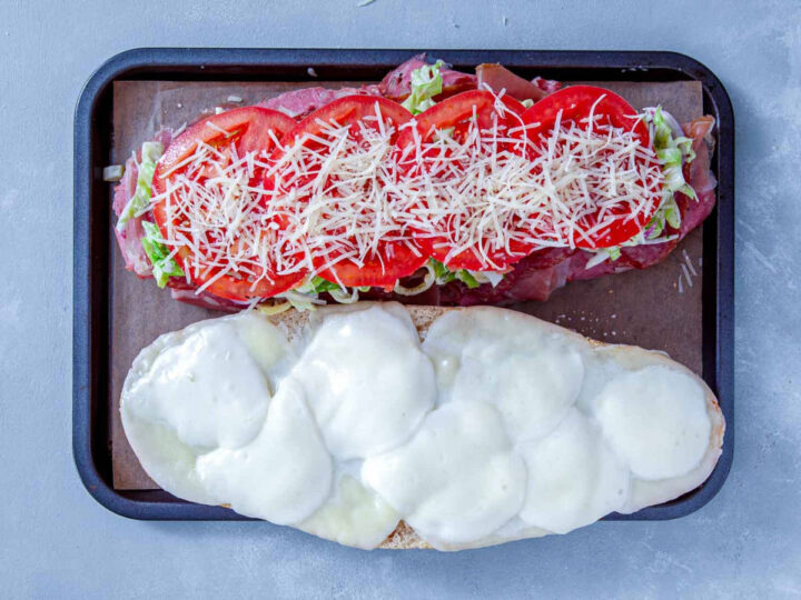 Italian grinder topped with sliced tomatoes and shredded cheese over lettuce and deli meats on a baking tray.