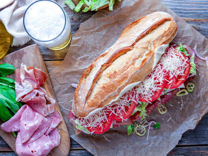 Italian grinder sandwich with deli meats, melted cheese, lettuce, tomato, and onions on parchment paper.