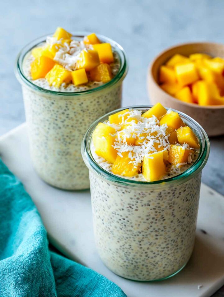 Two small glass jars filled with creamy off-white chia pudding, topped with bright yellow mango cubes and a light sprinkle of shredded coconut, set on a white marble surface with a teal napkin and a small bowl of extra mango in the background.