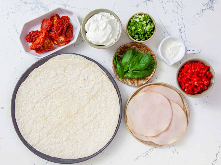 Ingredients for turkey pinwheels including tortillas, sliced turkey, cream cheese, sun-dried tomatoes, basil, and green onions.