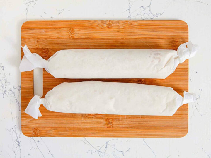 Tortilla rolls wrapped tightly in parchment paper, resting on a wooden cutting board.