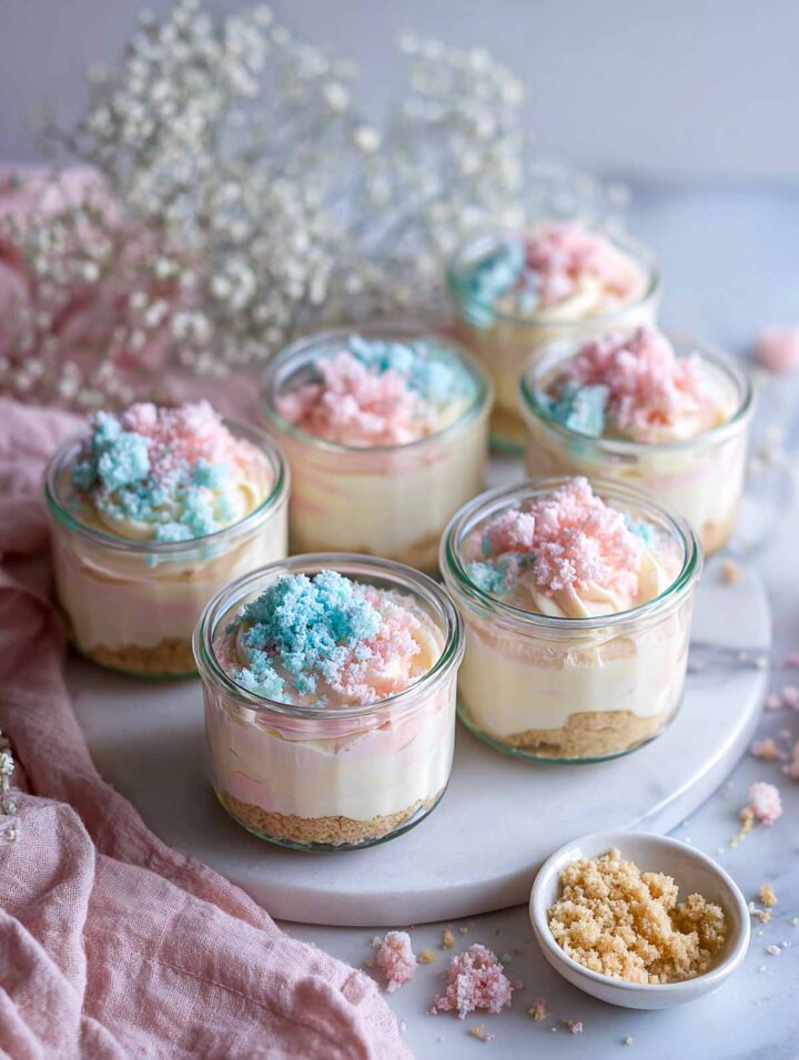Vanilla pudding cups layered with cookie crumbs and pastel crumble topping on a marble surface.