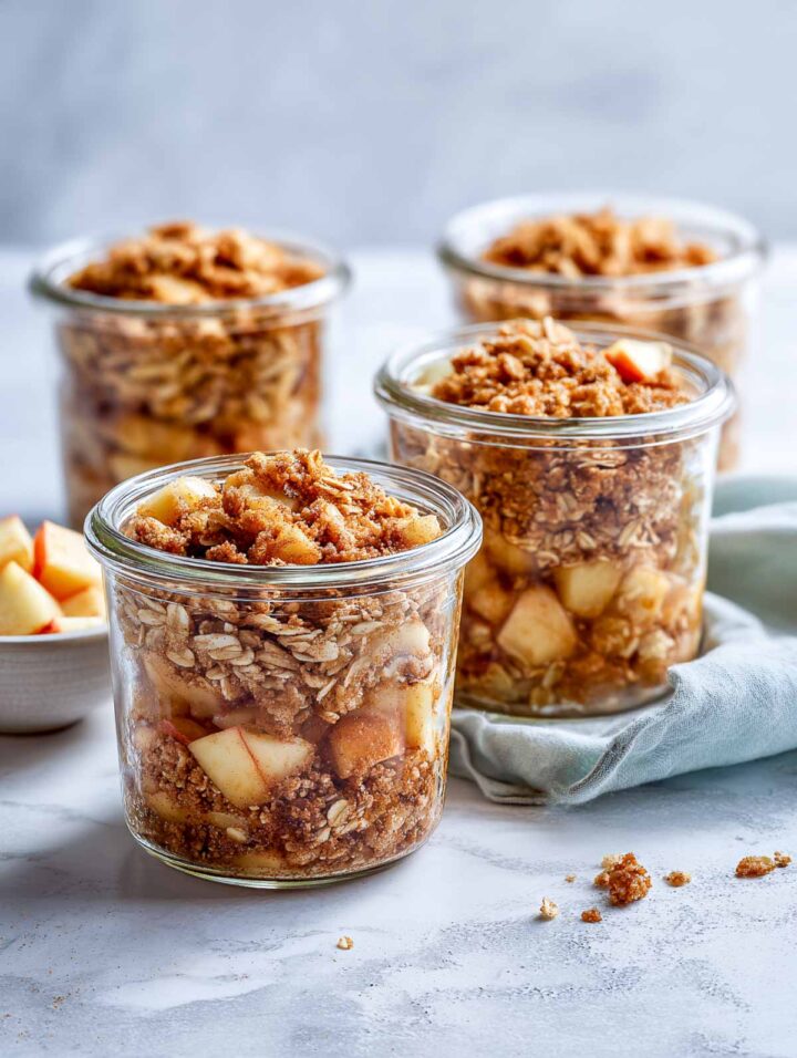 Apple crisp jars filled nearly to the rim with soft cinnamon-spiced apple pieces and a crumbly oat topping. The jars are arranged closely on a white marble surface with a soft sage napkin underneath. 