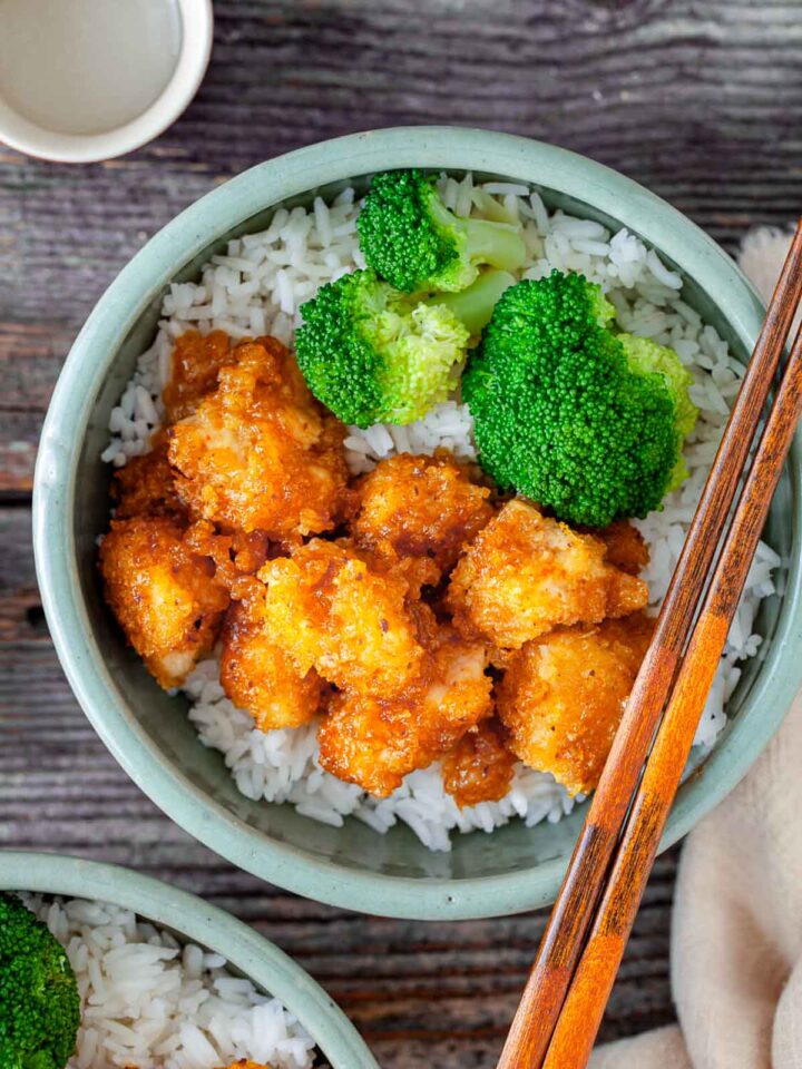 Bowl of orange chicken served over white rice with steamed broccoli and chopsticks.
