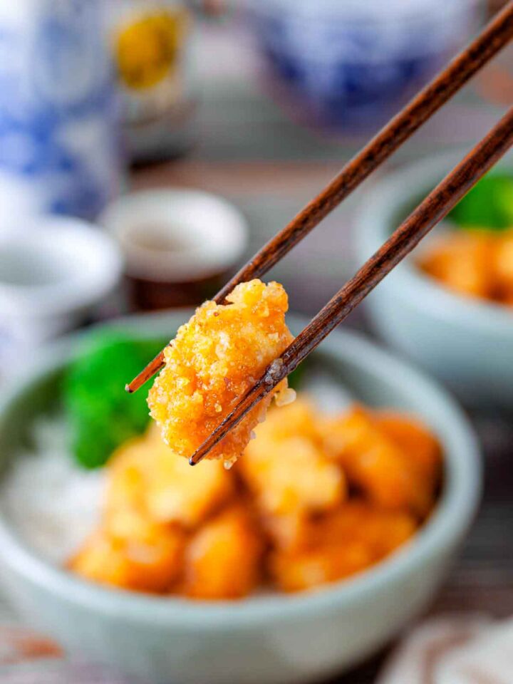Chopsticks holding a piece of crispy orange chicken above a bowl of rice and broccoli.